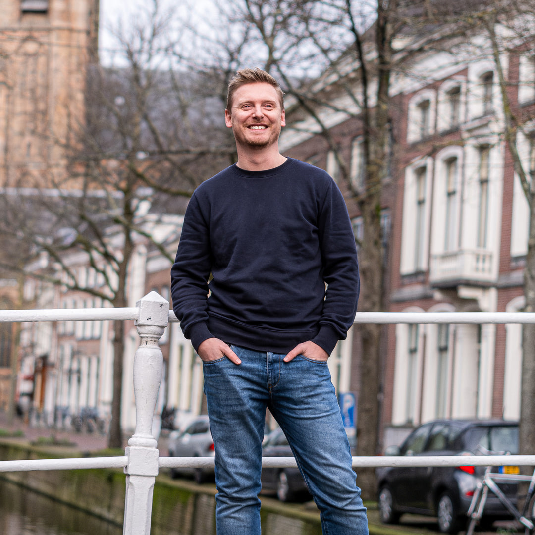 Man in Delft Denim jeans en een donkerblauwe trui poseert op een brug in Delft. Stijlvolle, duurzame heren spijkerbroek met perfecte pasvorm en tijdloos design.