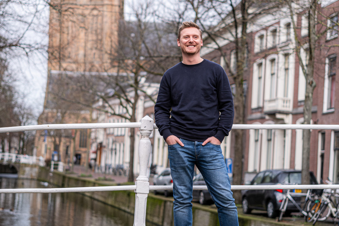 Man in Delft Denim jeans en een donkerblauwe trui poseert op een brug in Delft. Stijlvolle, duurzame heren spijkerbroek met perfecte pasvorm en tijdloos design.